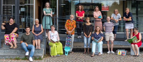 Das Bibliotheksteam präsentiert sich vor dem Eingang gegenüber der Reithalle. In den Händen halten sie verschiedene Medien vom Skateboard bis zum Buch.s