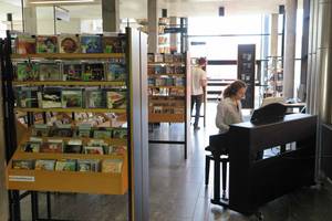 Die Musikbibliothek der Stadtbibliothek Offenburg wird gezeigt mit E-Piano und CD-Regalen. Eine Person sitzt am E-Piano, eine weitere Person wählt aus den Musik-CDs am Regal aus.