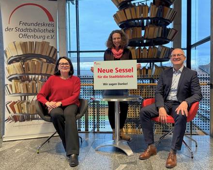 Quelle: ©Stadtbibliothek Offenburg Vor dem Flüsterturm der Stadtbibliothek sitzen die Freundeskreisvorsitzende und ein Vertreter einer Bank in den neuen Sesseln. Die Leiterin der Stadtbibliothek steht in der Mitte und präsentiert ein Plakat mit einem Dankeschön.