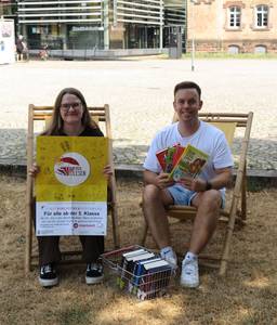 Lea und Jan präsentieren das Plakat der Aktion HEISS AUF LESEN in Liegestühlen sitzend vor der Stadtbibliothek Offenburg.