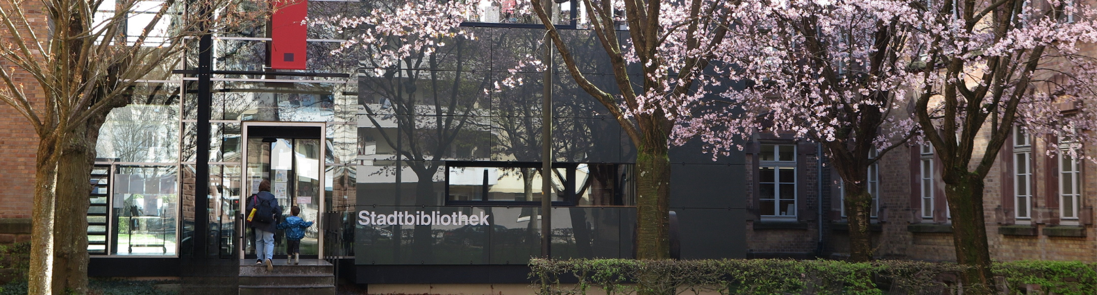Frau mit Kind betritt die Stadtbibliothek von der Weingartenstraße her. Die Szene ist frühlingshaft mit blühenden japanischen Kirschbäumen.