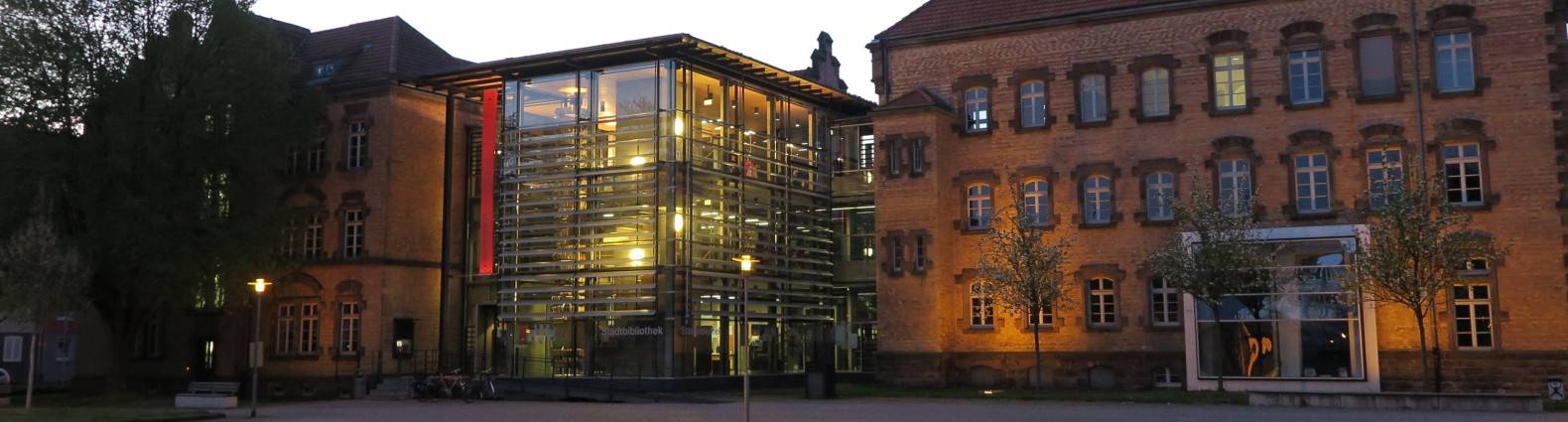 Die Stadtbibliothek im Dämmerlicht, gesehen vom Platz her, mit beleuchtetem Glaswürfel