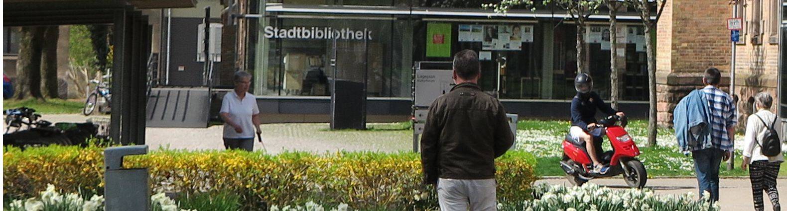 Seitlicher Blick auf die Stadtbibliothek. Im Vordergrund sind viele Menschen zu Fuß unterwegs. Ein Roller-Fahrer kreuzt.